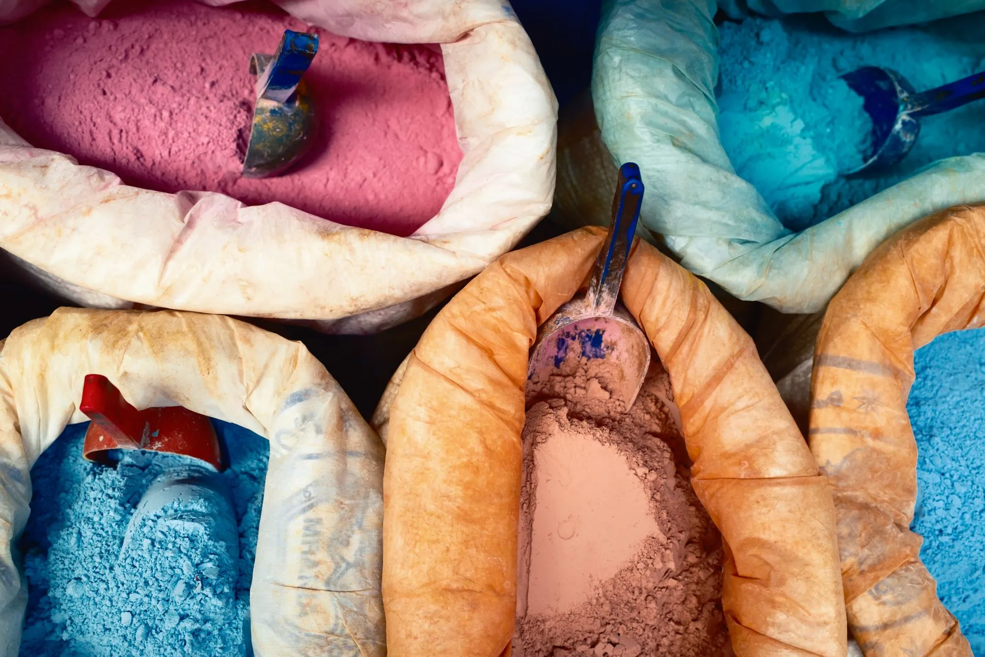 Open sacks of pigment powder in pink, blue, terracotta and pale green, each with a metal scoop